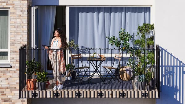 Une femme aux cheveux longs noirs debout sur le balcon d’un appartement avec des plantes en pot et une petite table avec deux chaises.
