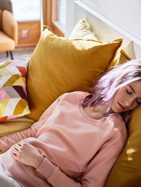 Une femme aux cheveux roses fait une rapide sieste dans son lit, appuyée sur un coussin avec housse GURLI jaune, assorti à la housse de couette.