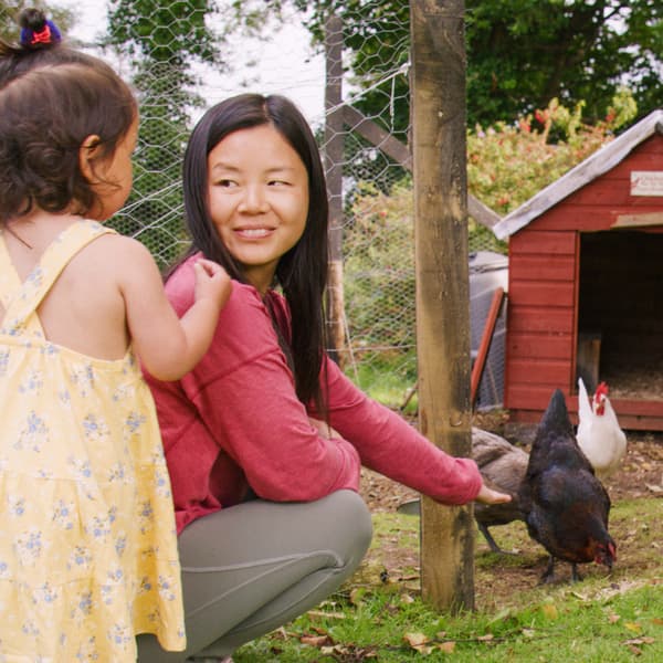 Une femme aux longs cheveux noirs et portant un chandail rouge nourrit ses poules avec sa fille.