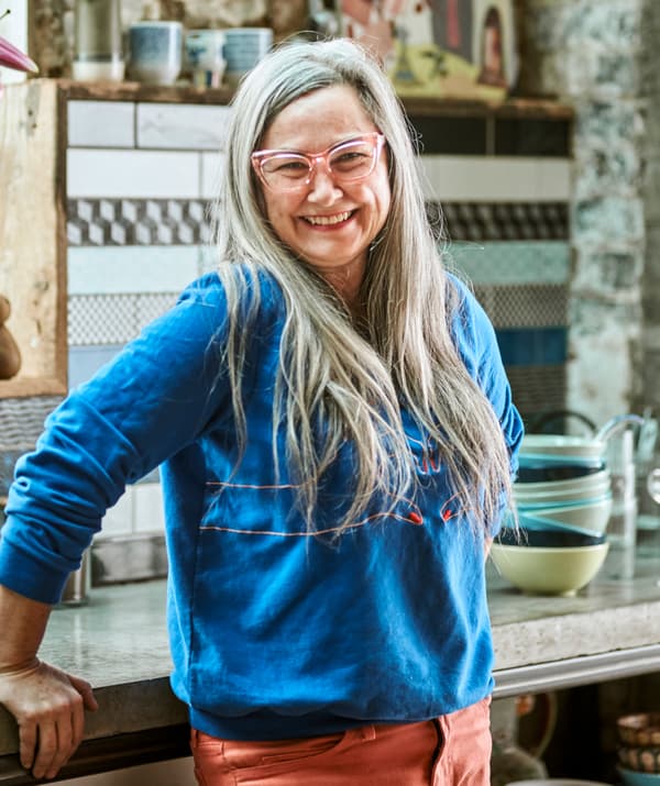 Une femme avec de longs cheveux gris et des lunettes à monture rose, une blouse bleue et un jeans rouge, souriant devant un plan de travail de cuisine.