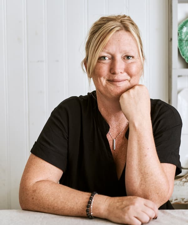 Une femme blonde souriante avec un t-shirt noir est assise à une table dans une pièce avec des murs en bois patinés.