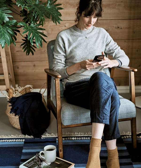 Une femme consulte son téléphone portable tout en étant assise dans un fauteuil avec un cadre en bois avec un rembourrage gris posé sur un tapis bleu.