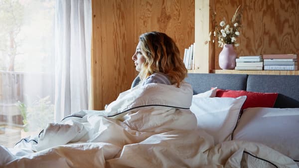 Une femme emmitouflée dans une couette blanche est assise sur un lit face à la fenêtre.