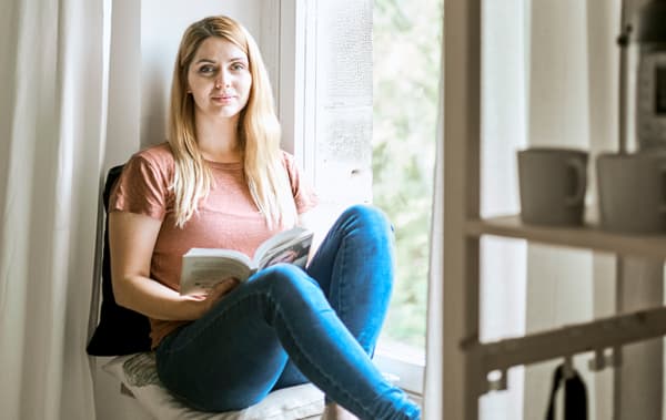 Une femme en jeans et T-shirt lit un livre assise sur un appui de fenêtre garni de coussins et d’un rideau blanc.