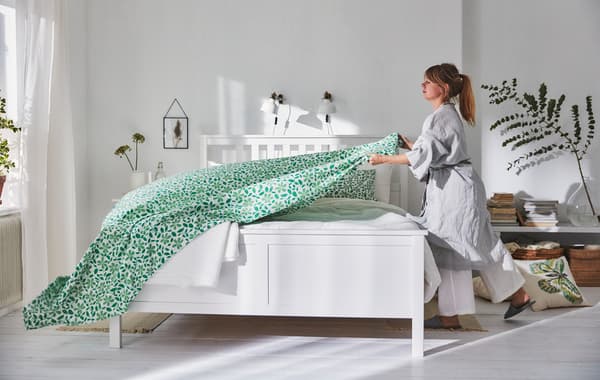 Une femme en robe de chambre gris clair recouvre un lit HEMNES d’un drap au motif de feuilles vertes.