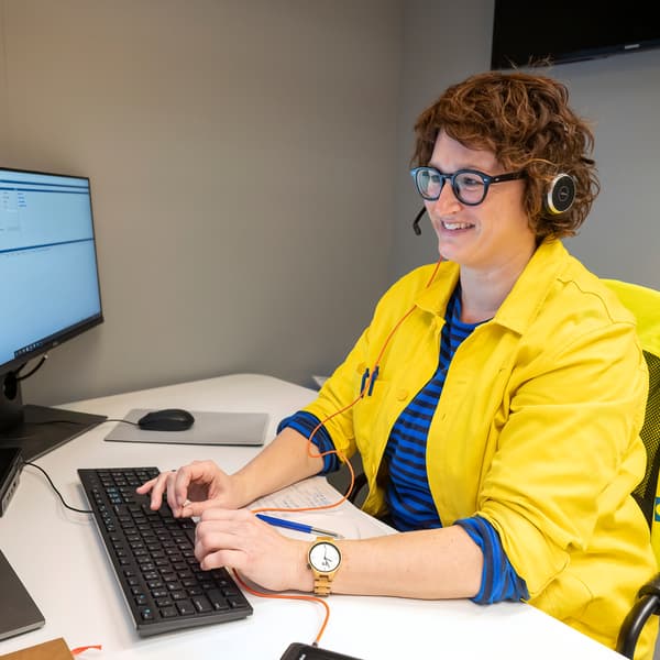 Une femme en tenue IKEA bleue et jaune, portant des lunettes et un casque audio, est assise à un bureau et regarde un écran en souriant.