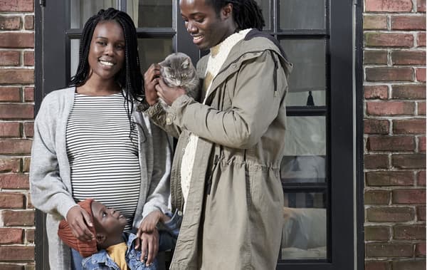 Une femme enceinte et un homme souriants, proches de la trentaine, devant un mur en briques, un petit garçon entre eux; l’homme a un chat dans les bras.
