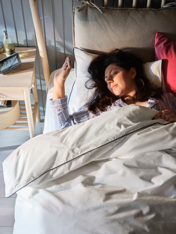 Une femme enveloppée dans une couette KUNGSBLOMMA protège ses yeux du soleil matinal qui pénètre dans la pièce.