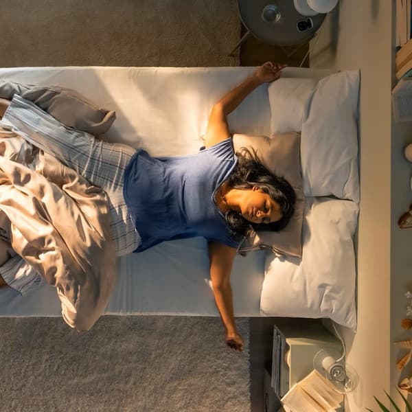 Une femme est allongée sur un lit bien fait, entourée de coussins moelleux et d'une couverture beige, dans une chambre confortable et chaleureusement éclairée.