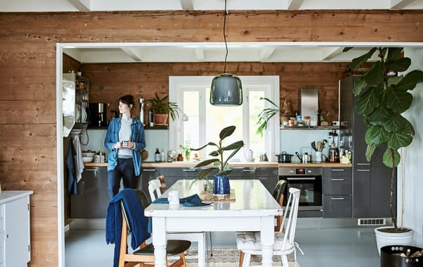 Une femme est appuyée contre une armoire gris brillant dans une cuisine qui s’ouvre sur une salle à manger avec une table et des chaises blanches.