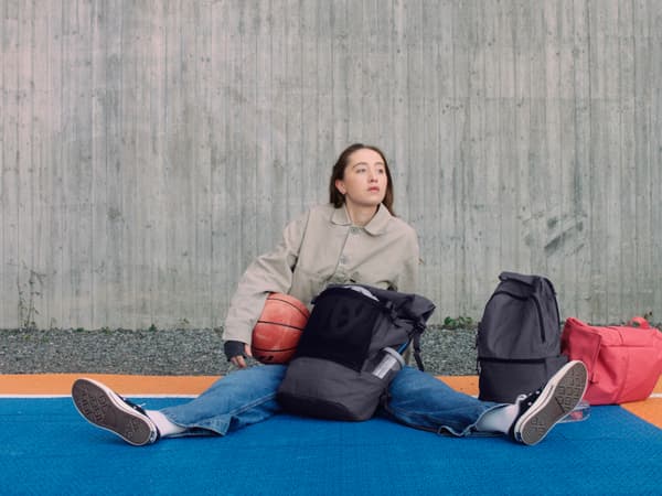 Une femme est assise sur le sol d'un terrain de sport, les jambes tendues, un ballon de basket à la main, et devant elle, sur le sol, un grand sac à dos de sport noir, dans la poche latérale duquel se trouve un gobelet et à côté duquel se trouvent deux autres sacs à dos