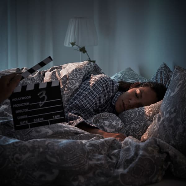 Une femme est endormie dans un lit face caméra alors qu’une main tient un clap de scène devant elle.