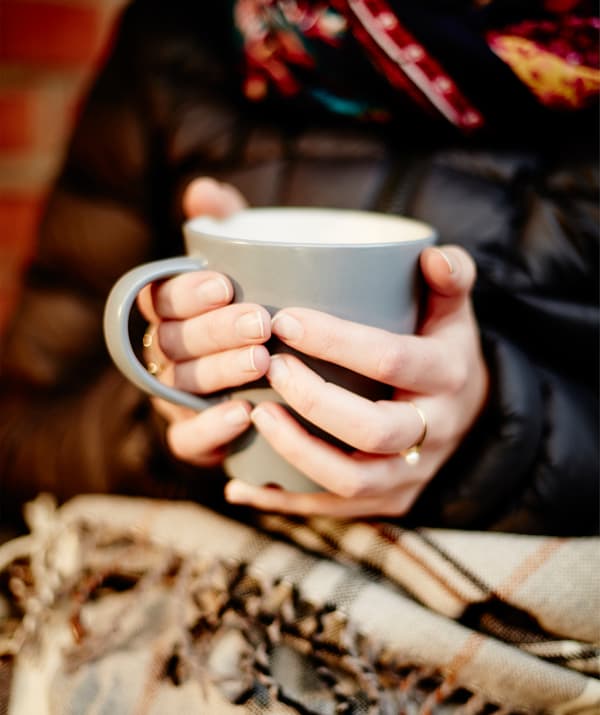 Une femme habillée chaudement avec un plaid sur les jambes et un mug fumant entre les deux mains.