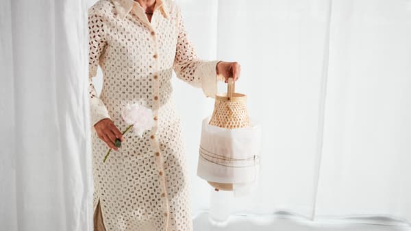 Une femme habillée d’une robe beige tenant une lampe de table MISTERHULT en bambou tressé, partiellement recouverte de papier.