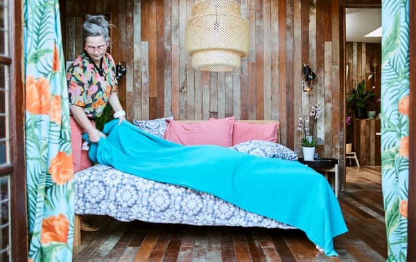 Une femme jette un plaid bleu sur un lit garni de draps à imprimé tie and dye, dans une chambre toute en lattes de bois, avec une grande suspension en matière tressée.