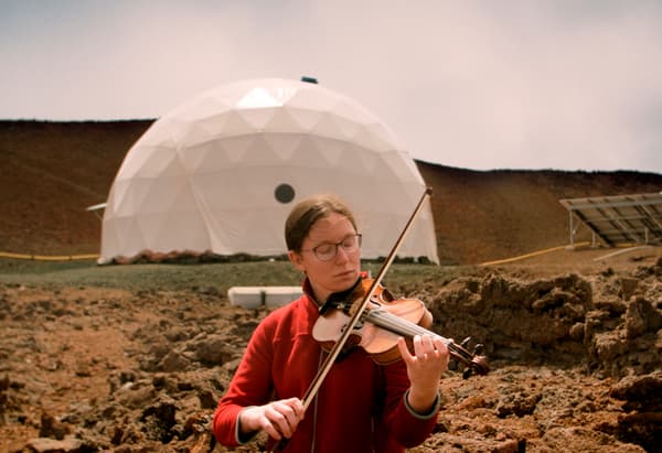 Une femme jouant du violon devant une structure fabriquée à la main dans un paysage aride.