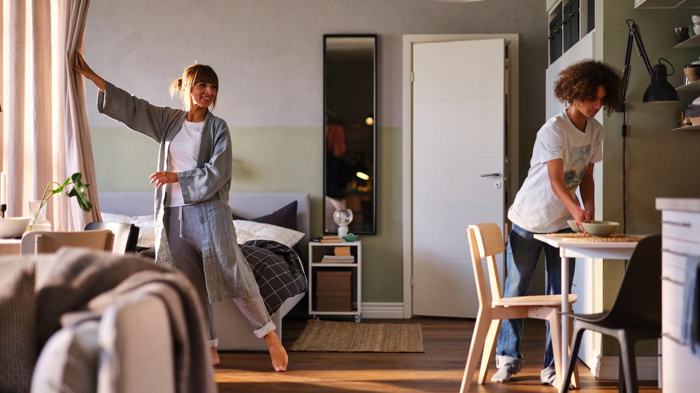 Une femme ouvre un rideau servant de séparation dans une pièce, révélant un lit matelassé GLADSTAD gris, tandis qu’un adolescent dresse la table du petit déjeuner.