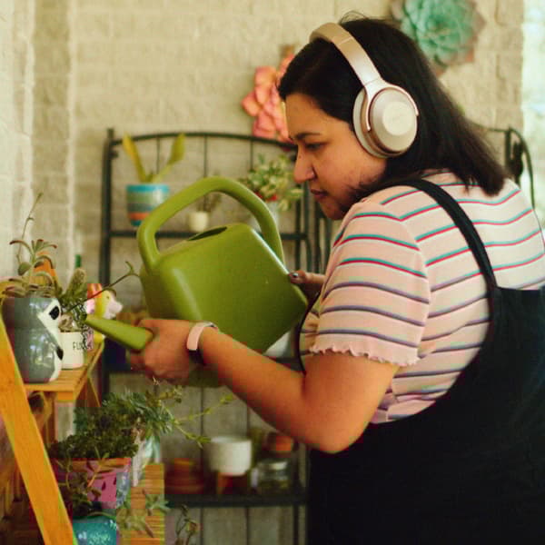 Une femme portant un casque d’écoute arrose des plantes, disposées dans des pots multicolores, avec un arrosoir vert.