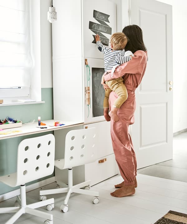 Une femme porte un enfant qui pointe du doigt des étiquettes tableau noir fixées sur une armoire blanche à côté d’un bureau avec deux chaises d’enfants à côté l’une de l’autre.
