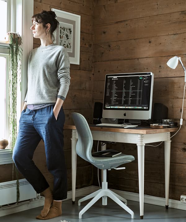 Une femme regarde par la fenêtre dans le coin d’une pièce transformée en bureau à domicile avec un bureau et une chaise pivotante.