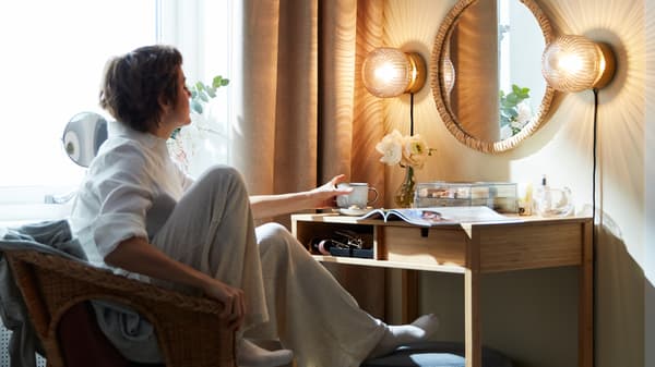 Une femme se repose dans un fauteuil et prend un gobelet sur une coiffeuse où il y a aussi un magazine, un vase à fleurs et une boîte transparente.