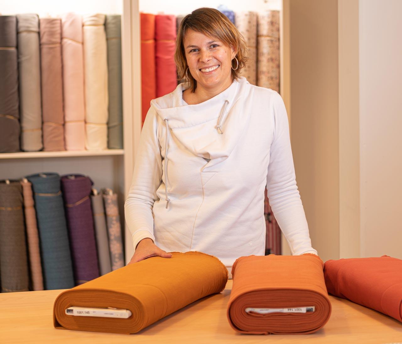Une femme se tient devant une étagère avec des rouleaux de tissu devant une table avec différents tissus.