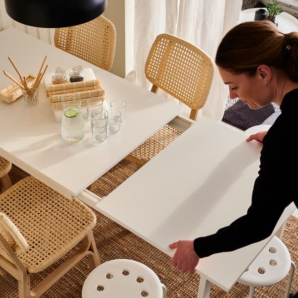 Une femme sortant la rallonge d’une table VANGSTA blanche sur un tapis en jute, avec des chaises en bambou clair et des tabourets blancs des deux côtés.