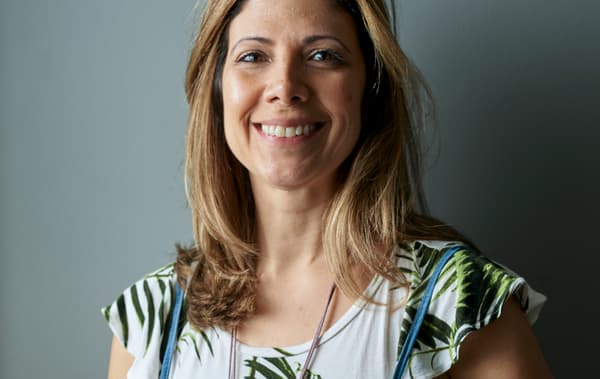 Une femme souriante aux cheveux marron méchés, debout devant un mur gris.