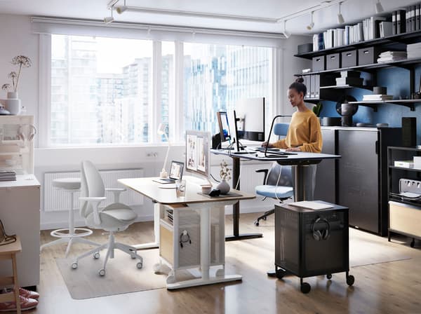 Une femme travaille à un bureau assis/debout BEKANT, à côté d’une grande fenêtre. Derrière elle, des dossiers sont rangés sur des tablettes ALGOT montées au mur.