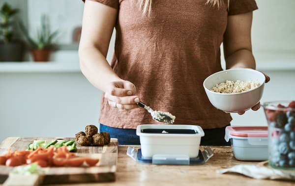 Une femme verse une cuillère de semoule d’un bol blanc dans un récipient en plastique. Sur une table en bois se trouve une planche à découper avec des légumes découpés.