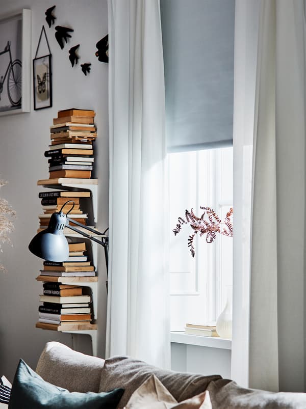 Une fenêtre avec des rideaux semi-transparents blancs, un store gris et un présentoir à livres vertical à côté d’une lampe de lecture.