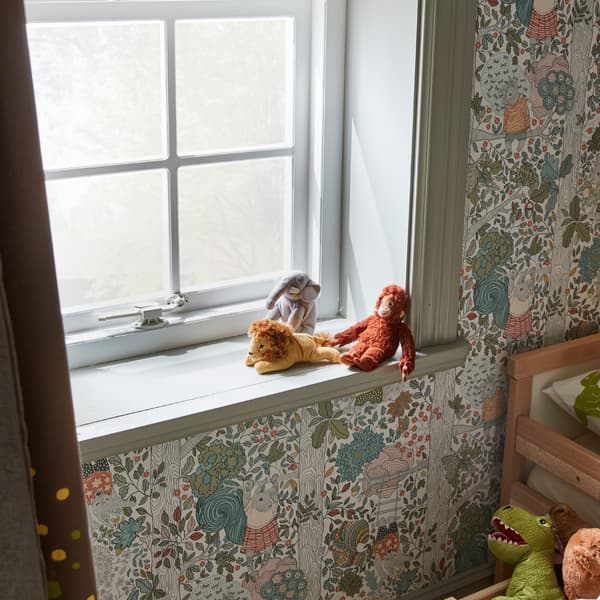 Une fenêtre équipée d’un bloque-fenêtre blanc, un appui de fenêtre vert sur lequel sont posées trois peluches, et des murs ornés de papier peint au motif de forêt.