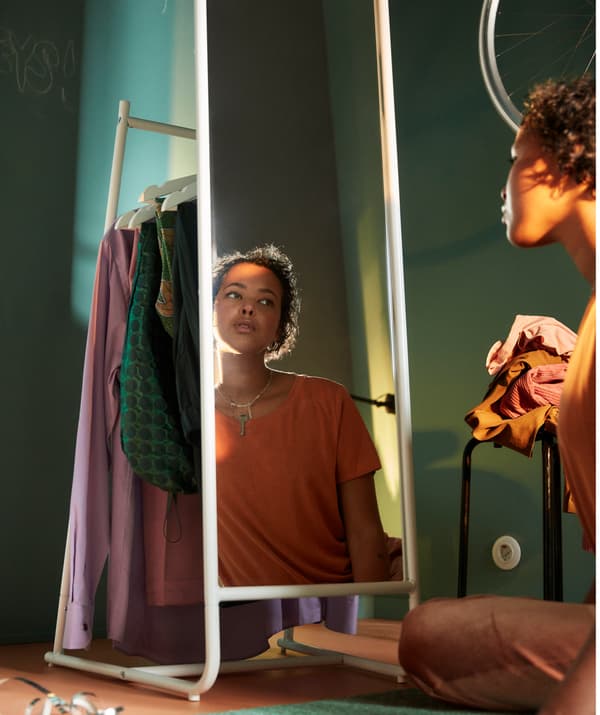 Une fille assise par terre se regardant dans le miroir avec une pile de vêtements posés sur un tabouret à ses côtés.