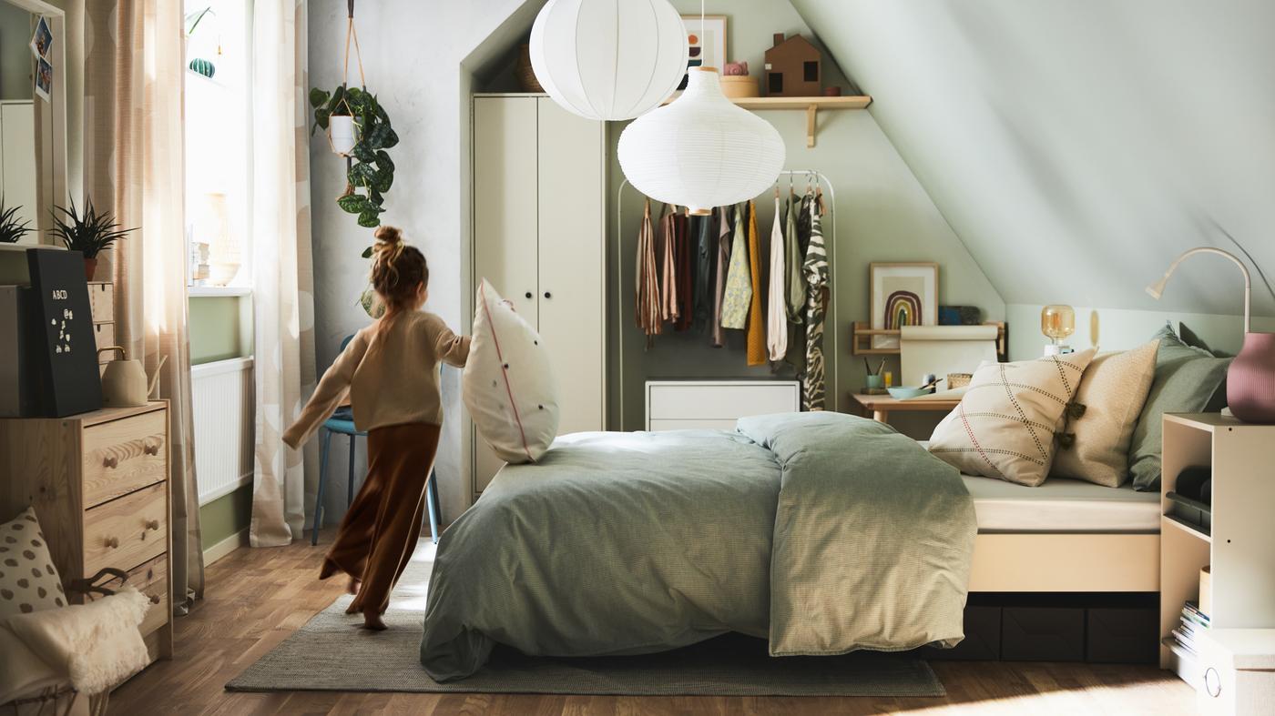 Une fille court devant un lit recouvert d'un linge de lit et de coussins aux rayures grises, ainsi que divers rangements et des suspensions blanches dans une petite chambre à coucher.