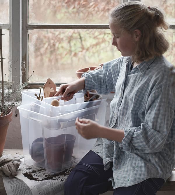 Une fille dans un décor de cabane de jardin occupée à retirer un séparateur avec des accessoires de jardin d’une boîte transparente SAMLA.