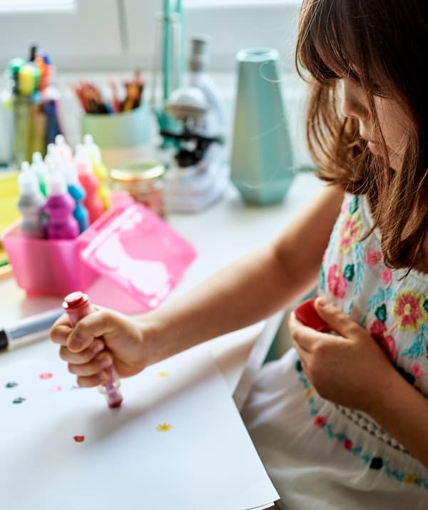 Une fillette assise à un bureau utilisant un feutre tampon rouge pour tamponner des motifs sur du papier, une boîte rose contenant des tubes de peinture à côté d’elle.