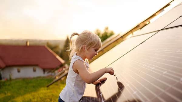 Une fillette blonde effleure un panneau solaire avec un brin d’herbe, sur le toit d’une maison à la campagne.