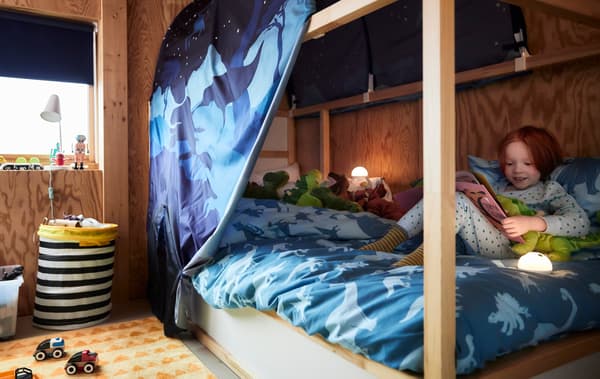 Une fillette lit un livre d’histoires en câlinant son dinosaure en peluche, assise sur son lit KURA, sous un ciel de lit au motif de dinosaures. 