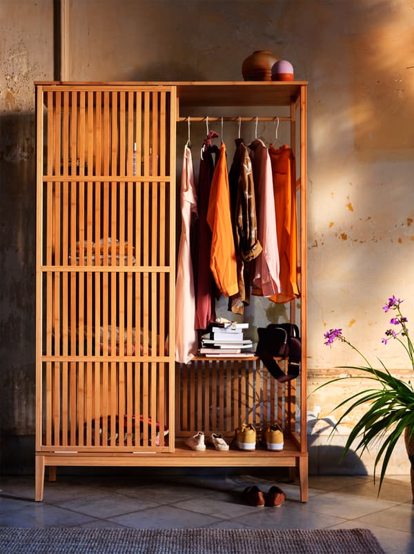 Une garde-robe ouverte en bois avec des portes coulissantes remplies de vêtements aux couleurs de l’automne, placée contre un mur jaune-orange.