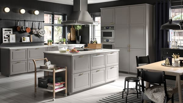 Une grande cuisine grise avec un îlot de cuisine au centre, flanqué de tabourets noirs, et une table de salle à manger en hêtre/blanc entourée de chaises noires.