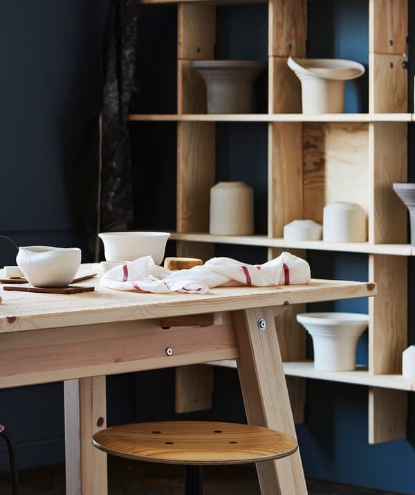 Une grande table en bois blond non traité dans une pièce peinte avec des couleurs riches; des pots disposés dans une grande étagère murale en bois blond à l’arrière-plan.