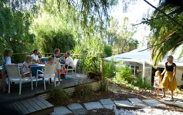 Une groupe de personnes assises autour d'une table sur une terrasse surélevée dans un jardin avec de la verdure et des dalles.