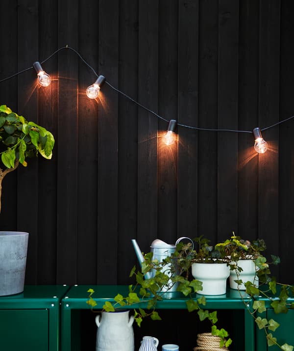 Une guirlande lumineuse accrochée à un mur brun foncé éclaire la terrasse et deux armoires vertes.