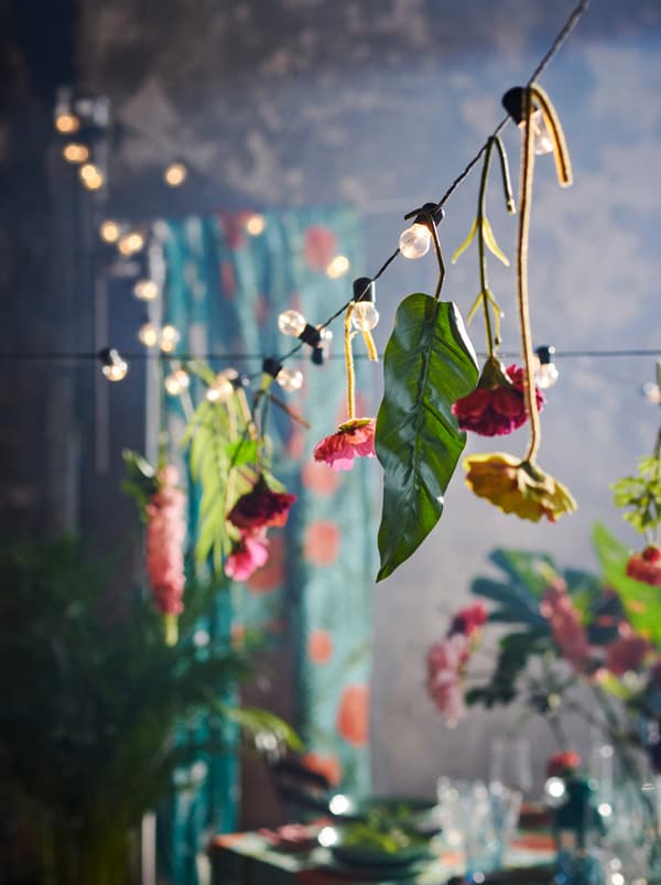 Une guirlande lumineuse BLÖTSNÖ avec des fleurs artificielles SMYCKA suspendues de-ci de-là, tendue au-dessus d’une table dressée et décorée.