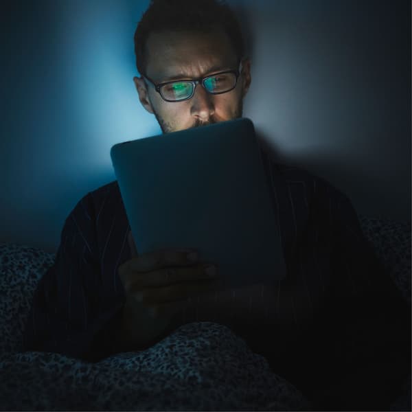 Une homme avec des lunettes regarde un iPad dans un lit. La lumière de l’écran se reflète sur son visage fatigué.
