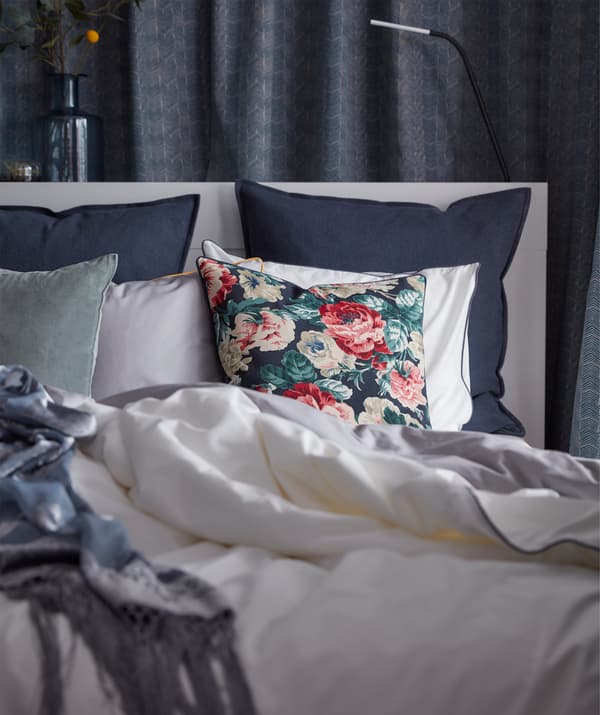 Une housse de coussin noire et multicolore IKEA LEIKNI avec un motif floral, déposée sur un lit avec d’autres coussins.