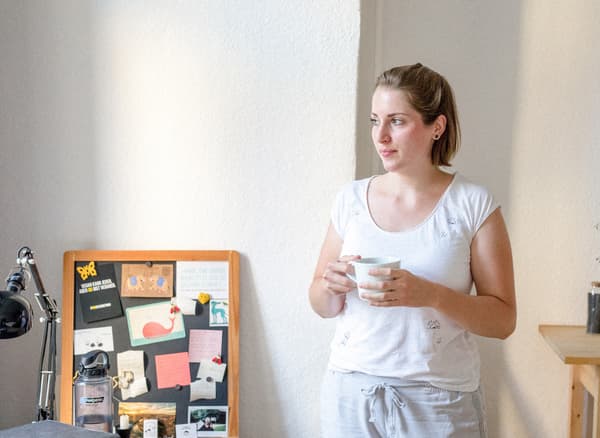 Une jeune femme chez elle, une tasse de thé à la main, qui regarde par la fenêtre.