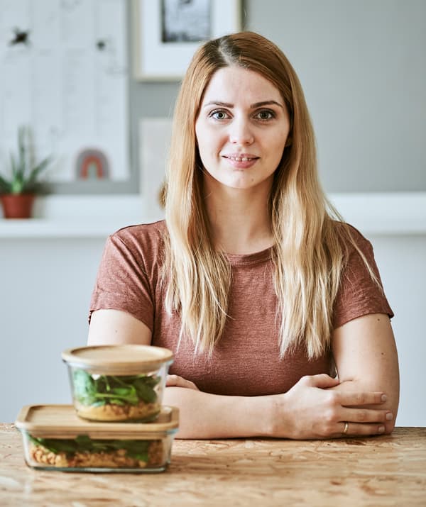 Une jeune femme est assise à une table en bois avec devant elle deux bocaux en verre avec un couvercle en bambou contenant un ragoût de lentilles et des épinards.