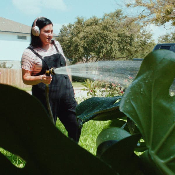 Une jeune femme portant une salopette et des écouteurs tient un boyau d'arrosage pour arroser une grande plante de jardin.