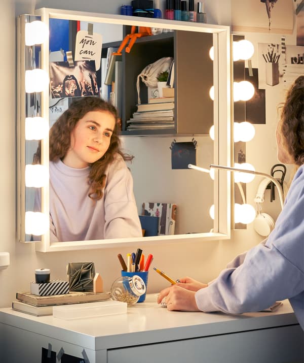 Une jeune femme se regarde dans un miroir éclairé et disposé au-dessus d’un bureau blanc.